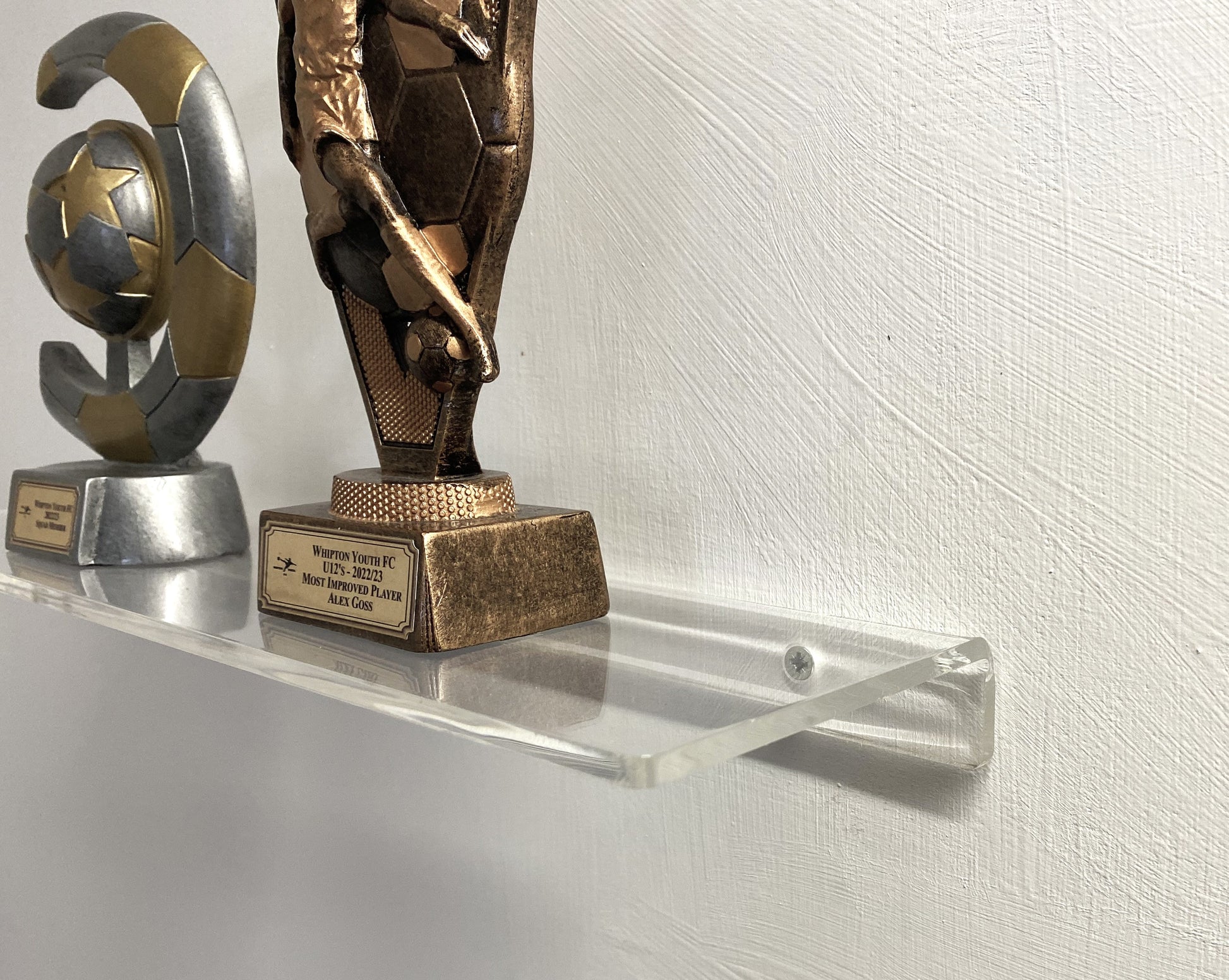 Two trophies on a clear shelf against a white wall